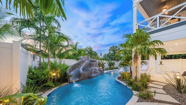 A swimming pool with a waterfall feature, surrounded by tropical plants and a modern building with a balcony. - 2 Homes–2 Pools/Hot Tubs–Steps from Siesta Beach (Sarasota)