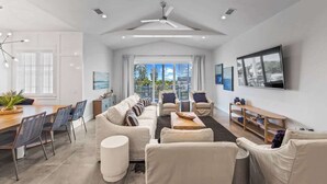 Un salon moderne comprenant un ensemble de canapés, un coin repas avec des chaises, un ventilateur de plafond et un téléviseur fixé au mur.