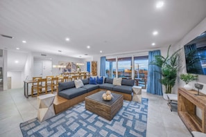 A modern living room with a sectional sofa, dining area, and large windows offering a view. - New - Dual-home luxury, Rooftop Decks, Sleeps 32 (Siesta Key)