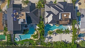 Vista aerea di un resort con diversi edifici, una grande piscina centrale e palme circostanti.