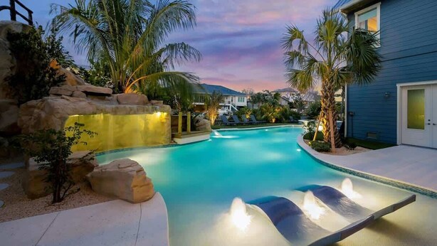 Una piscina con cascada, rodeada de palmeras y una casa.
