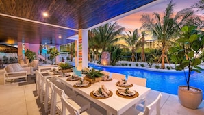 Un espacio para comer al aire libre con piscina, palmeras y vistas al atardecer.