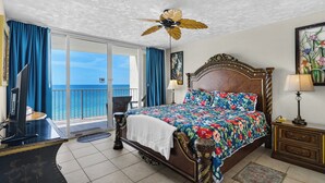 Habitación de hotel con una cama grande, ventilador de techo, vista al océano y balcón con cortinas azules.