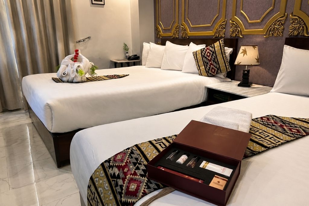 A hotel room with two beds, a decorative headboard, a bedside table with a lamp, and a tray with toiletries.