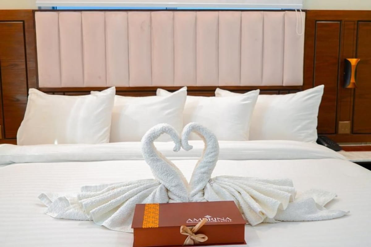 Una habitación de hotel con cama, almohadas, una toalla dispuesta en forma de corazón y una caja de regalo.