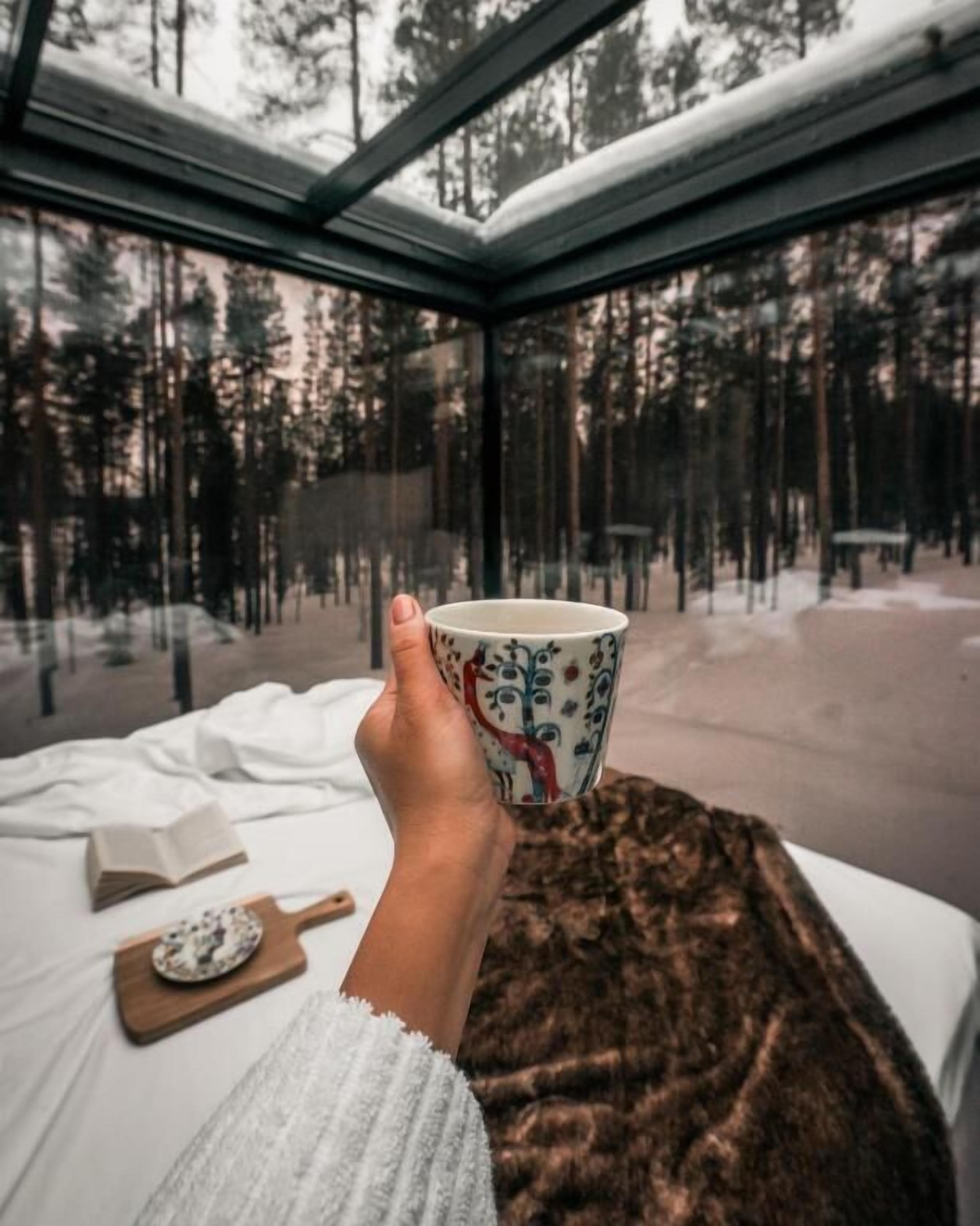 Eine Hand hält eine Tasse, ein Buch und ein Holzbrett auf einem Bett, mit Blick auf einen verschneiten Wald durch ein Fenster.