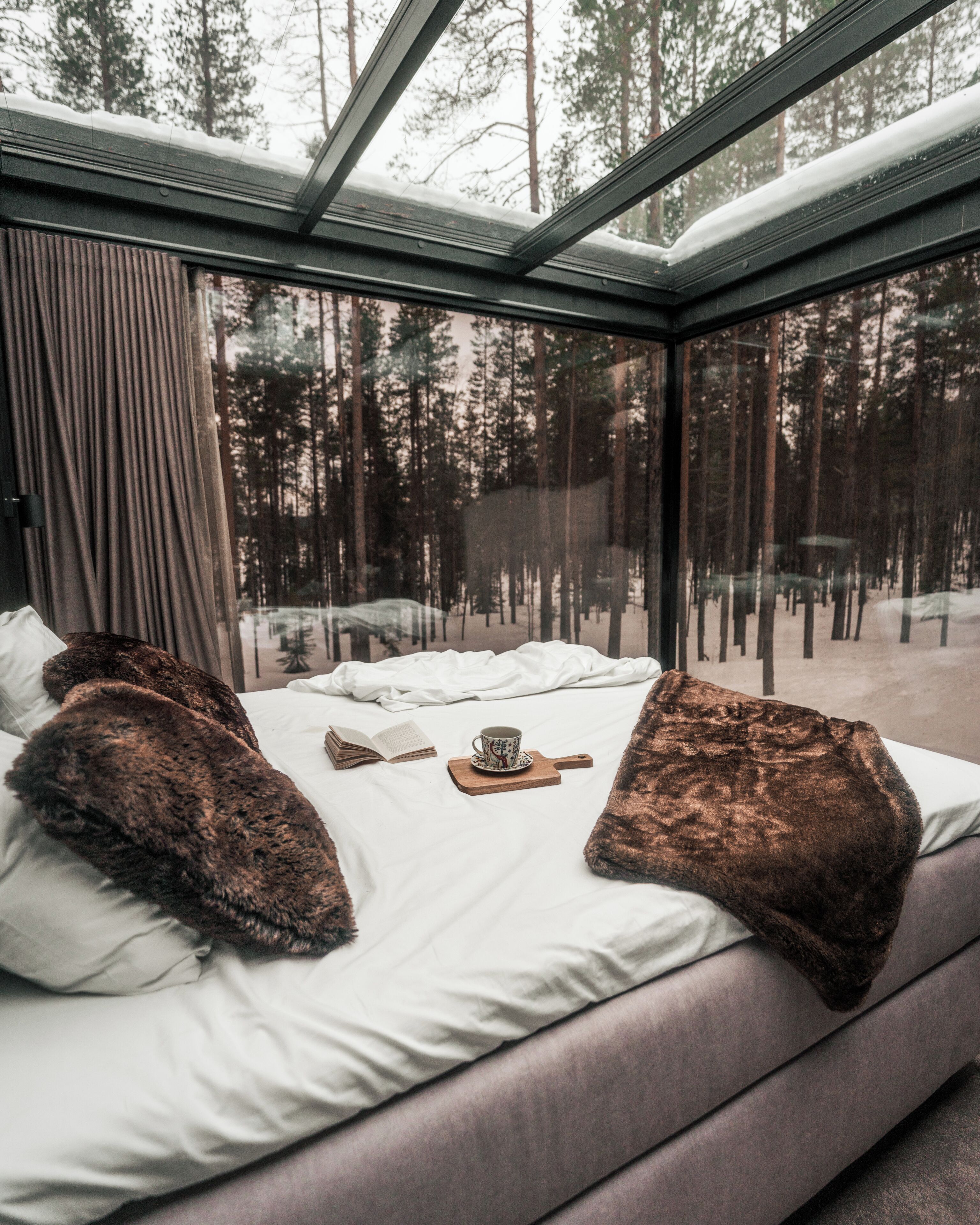 Une chambre avec une grande fenêtre, un lit avec des oreillers, une tasse et un livre.