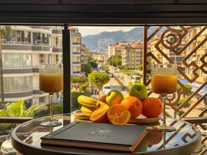 Ein Tisch mit zwei Gläsern Saft, Obst und einem Buch auf einem Balkon mit Blick auf eine Stadtlandschaft.
