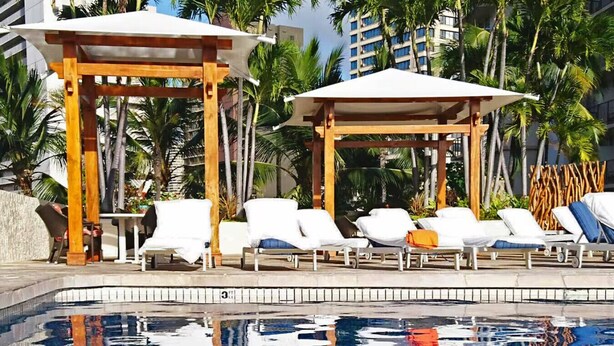 Zona junto a la piscina con sillones de descanso y sombrillas, rodeada de palmeras y un edificio al fondo.