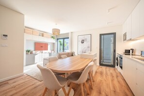 A modern kitchen with a dining area, featuring a wooden table and chairs, a built-in oven, and a large window with blinds. - GuestReady - Maleo Living Apartments - 7C2 (Lisbon)