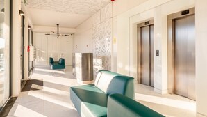 Um lobby moderno com balcão de recepção, dois elevadores e área de estar com cadeiras cor turquesa.