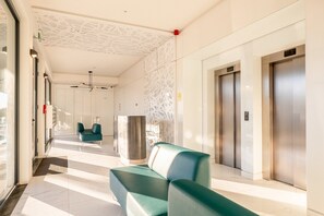 Eine moderne Lobby mit einer Rezeption, zwei Aufzügen und einer Sitzecke mit türkisfarbenen Stühlen.