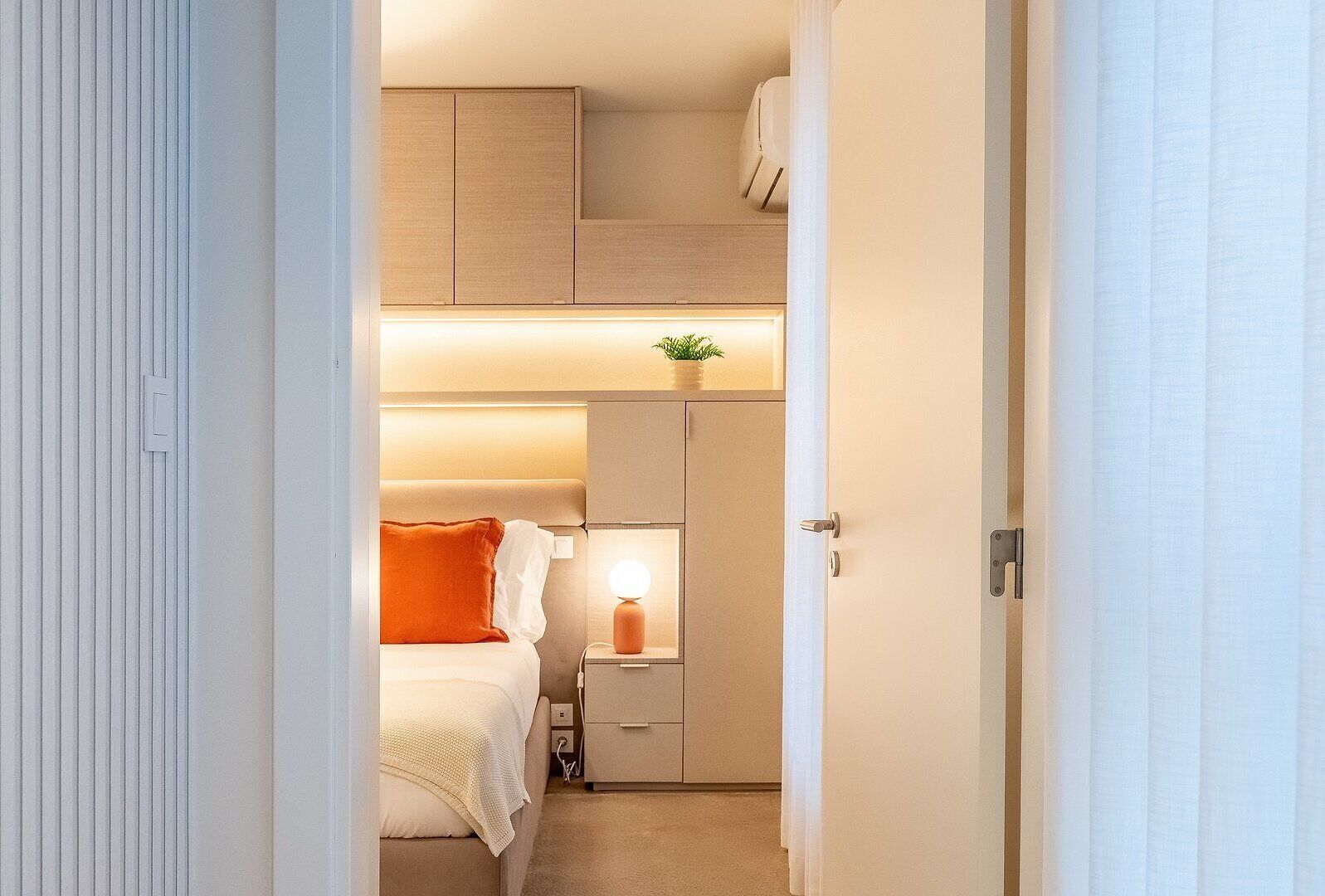 Un pasillo estrecho y bien iluminado que conduce a un dormitorio con cama, mesita de noche y una pequeña planta.