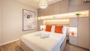 Quarto de hotel moderno com uma cama grande, mesas de cabeceira com luminárias, cabeceira com iluminação integrada, um quadro na parede e uma pequena planta na mesa de cabeceira.