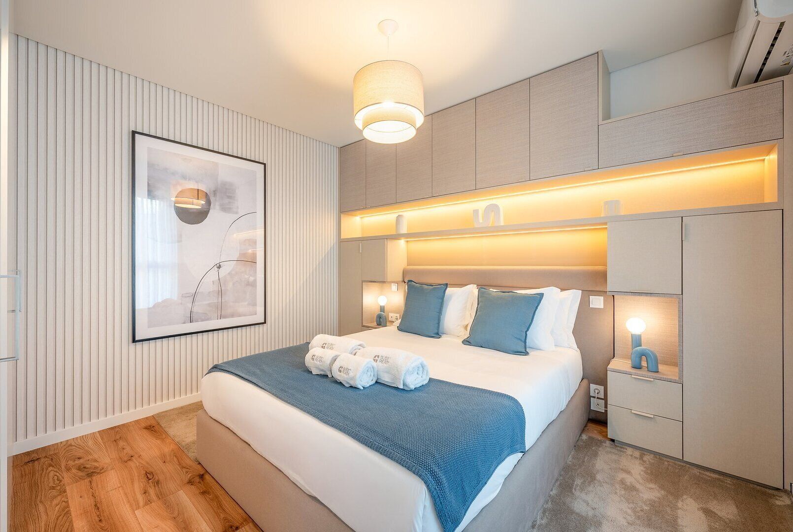 Une chambre à coucher moderne, avec un lit, des tables de chevet, un luminaire fixé au mur et une œuvre d’art encadrée.