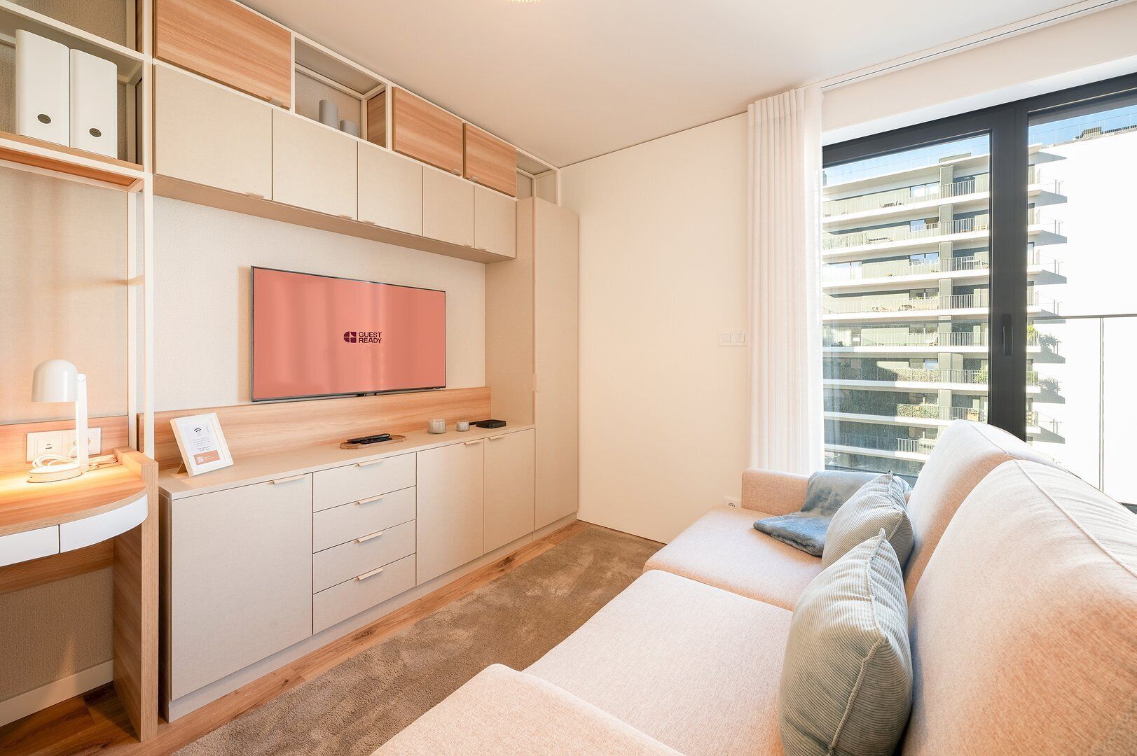 Een moderne hotelkamer met een kitchenette, een flatscreen-tv, een bank met kussens en een groot raam met uitzicht op de stad.
