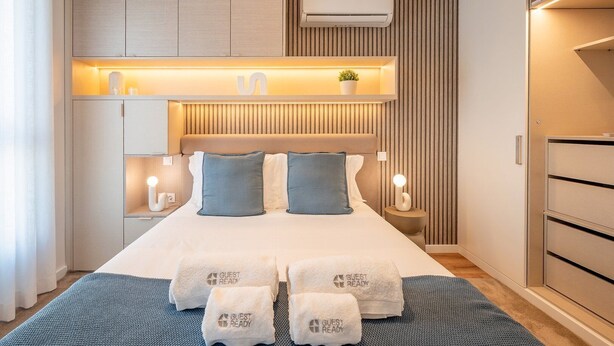 Ein modernes Hotelzimmer mit Bett, Nachttischlampen, einem Nachttisch, einer Wand-Klimaanlage und einem eingebauten Regal mit Pflanze.