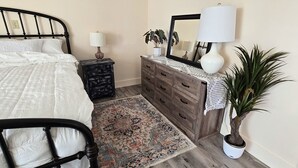 Ein Schlafzimmer mit Bett, Schminktisch, Nachttisch, Lampe, Spiegel, Teppich und einer Zimmerpflanze.