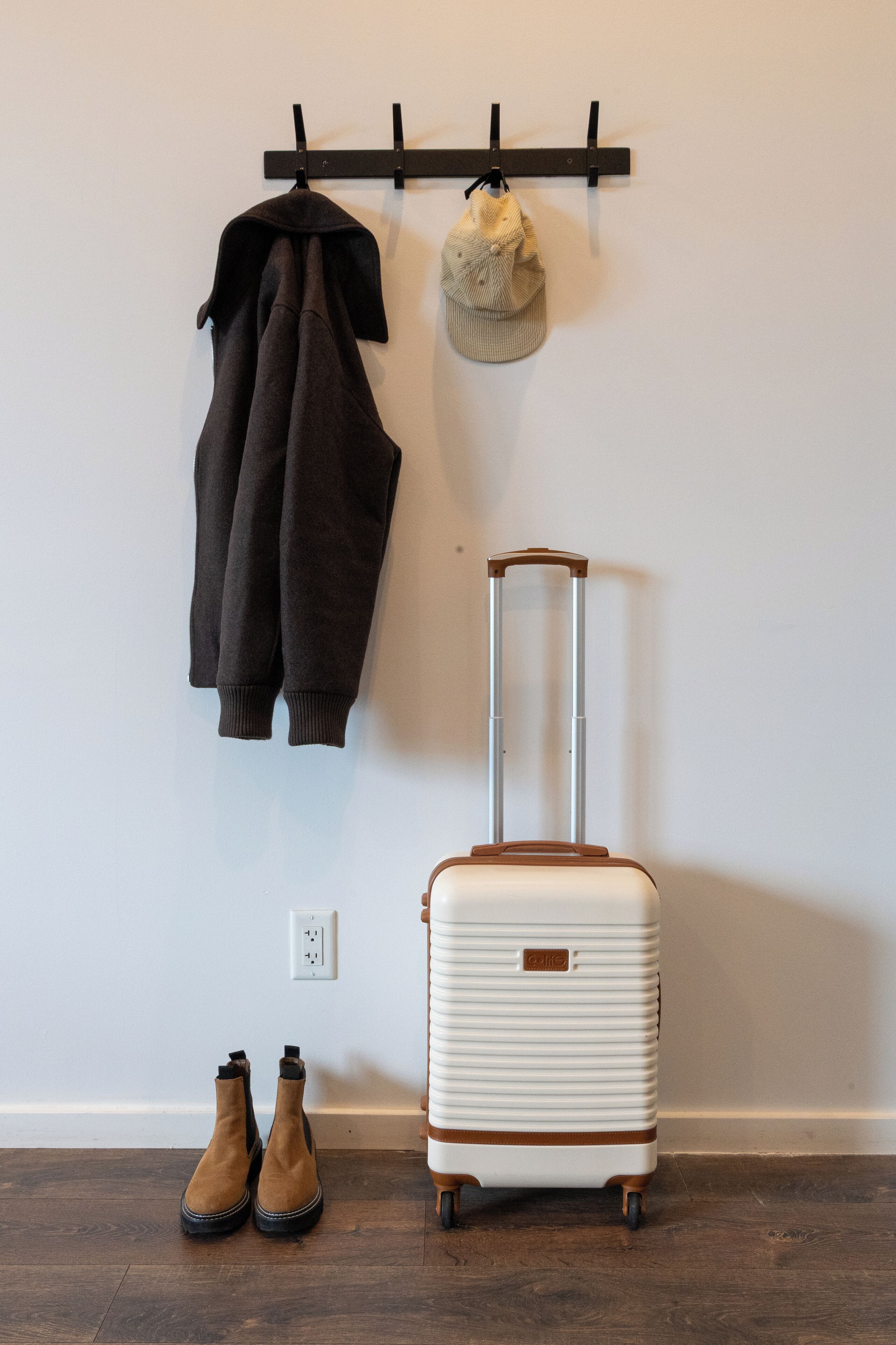 A room with a white suitcase, a pair of brown boots, a black jacket, and a beige hat hanging on a black wall-mounted rack.