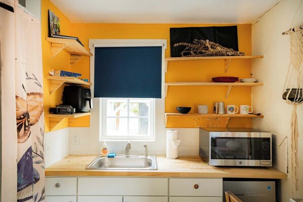 Cocina compacta con lavamanos, microondas y repisas con vajilla y libros.