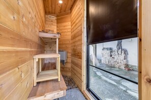 Interior d'una cabana de fusta amb una taula de fusta petita, un graó que condueix a una zona en alt i una finestra amb vista a una pila de maons a l'exterior.