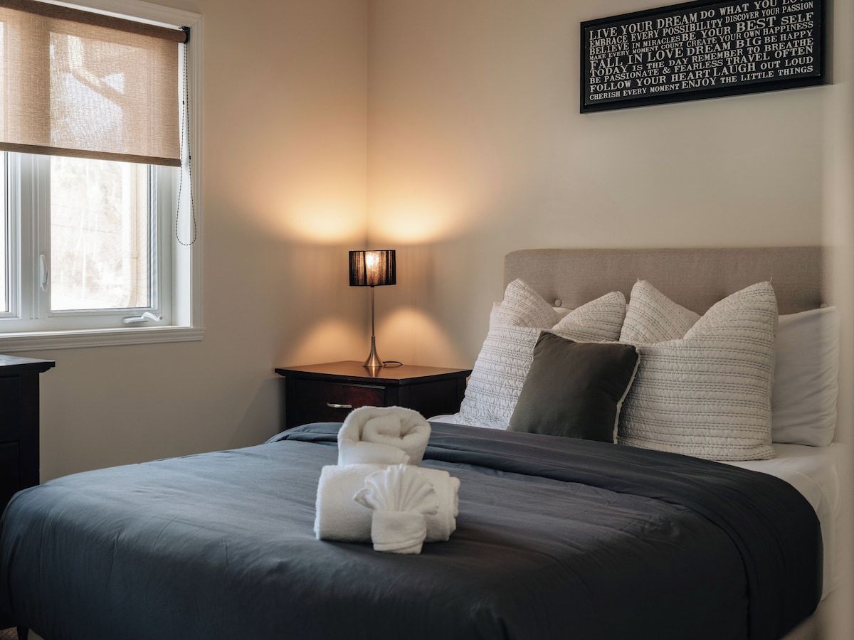A neatly made bed with a dark bedspread and two rolled towels, a bedside lamp, and a wall with motivational text.