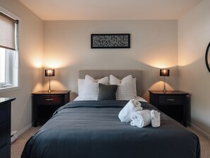 Un lit bien fait, avec une couvre-lit et des oreillers aux couleurs sombres, deux tables de chevet avec des lampes et une citation inspirante accrochée au mur.