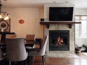 Una sala de estar con chimenea, una zona de comedor con sillas, un televisor de pantalla plana sobre la chimenea y un reloj en la pared.