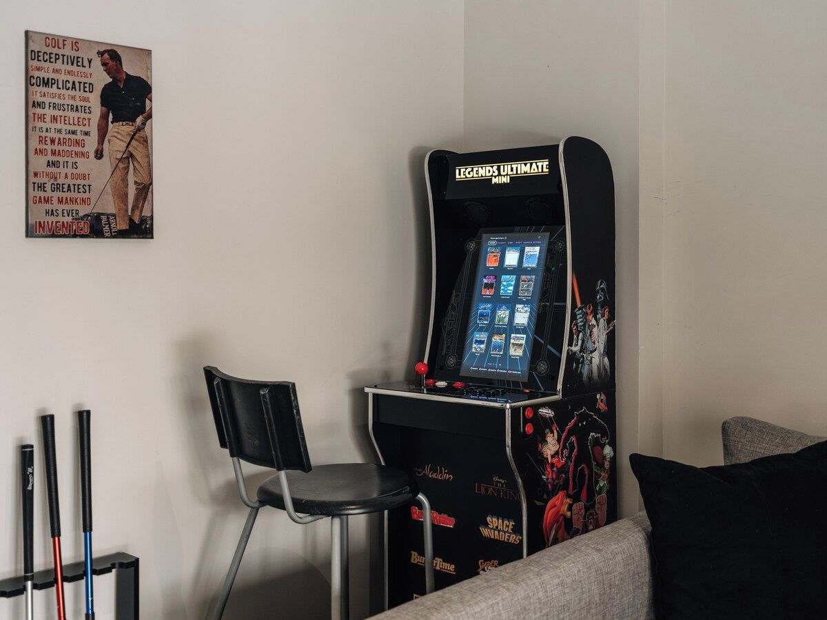 A room with a Legends Ultimate Mini arcade machine, a black chair, and a couch with a pillow.