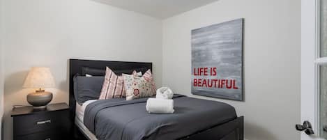 Un dormitorio con cama, mesita de noche, lámpara y una frase motivacional en la pared.