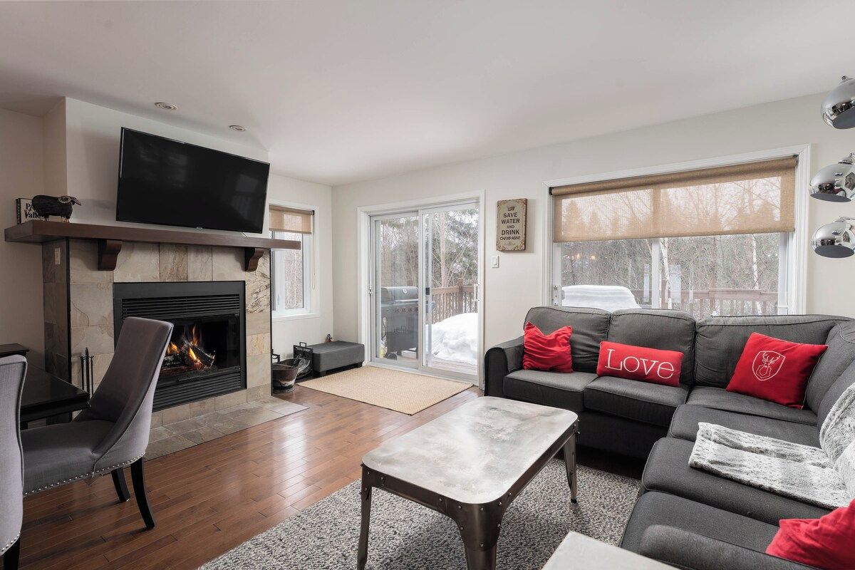 A living room with a fireplace, a television, a sofa with red cushions, and a large window.