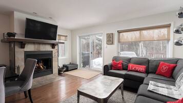 A living room with a fireplace, a television, a sofa with red cushions, and a large window.