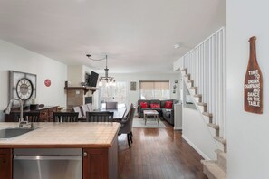 Cocina moderna con isla central, comedor con sillas, escalera y sala de estar con sofá y cojines decorativos.