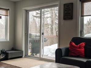 A living room with a sliding glass door leading to a deck, a black sofa with a red pillow, and a barbecue grill outside. - MiniPutt ~ Arcade ~ Pool ~ Playground ~ Golf & Ski (Mont-Tremblant)