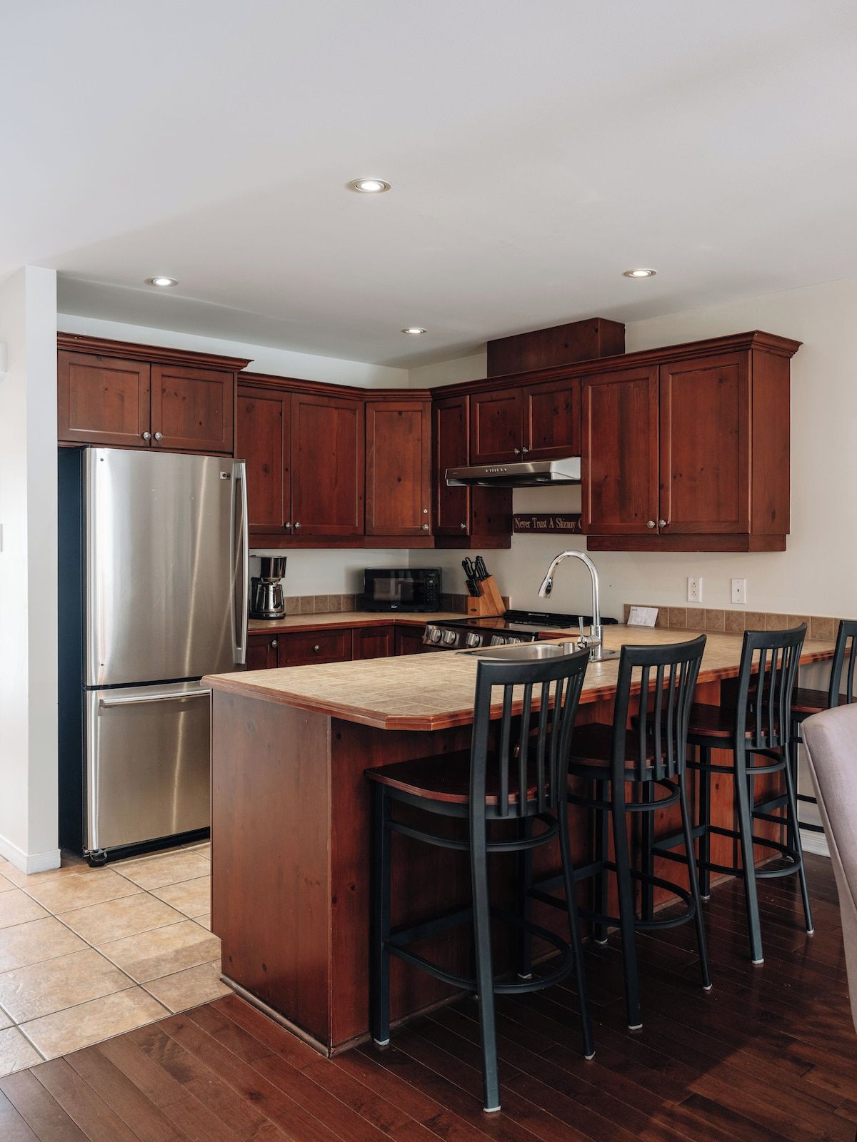 A modern kitchen with wooden cabinets, a stainless steel refrigerator, a central island with bar stools, and a built-in oven.