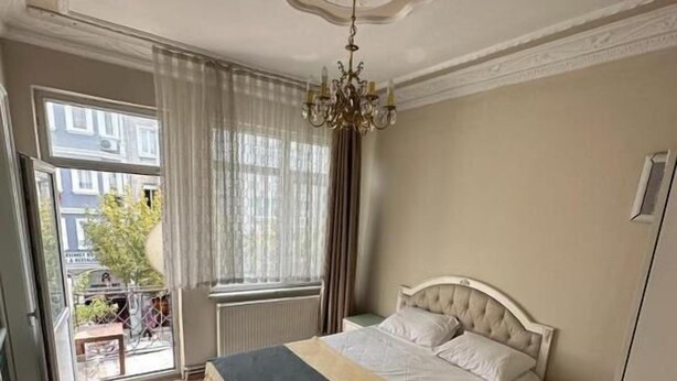 Une chambre à coucher avec un lit, une table de chevet, un lustre, une fenêtre avec des rideaux et une vue sur l’extérieur.