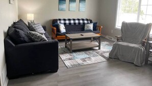 Sala de estar com um sofá preto, uma poltrona cinza, uma mesa de centro de madeira e um tapete estampado. Há três quadros abstratos na parede e uma janela com persianas.
