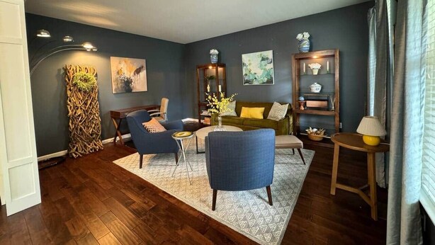 A living room with a sofa, two armchairs, a coffee table, and a side table with a lamp. There is a decorative plant, a painting, and a bookshelf with books and decorative items. - The Battlefield Getaway (Monroe)