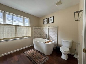 A bathroom with a freestanding bathtub, a toilet, and a window with curtains. - The Battlefield Getaway (Monroe)