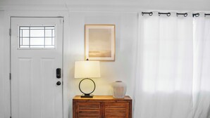 Une porte blanche avec une fenêtre, un meuble en bois orné d’une lampe et d’un vase, et un rideau blanc.