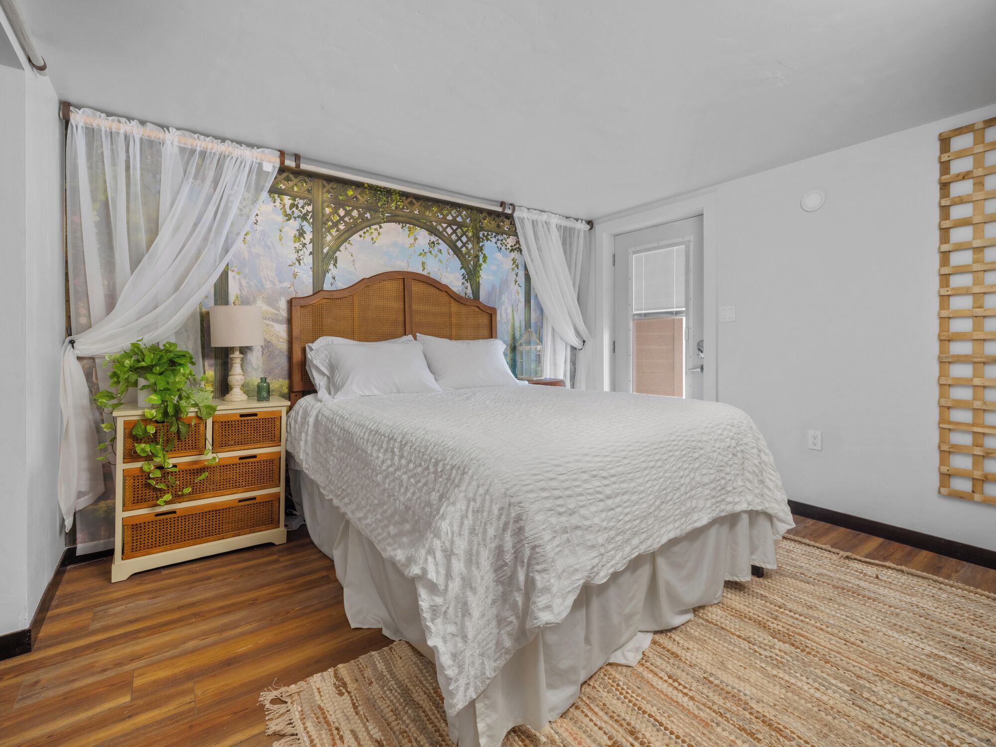 Une chambre à coucher avec une tête de lit en bois, du linge de lit blanc, une table de chevet en osier avec une lampe et un tapis tissé.
