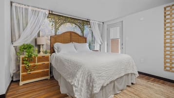 Une chambre à coucher avec une tête de lit en bois, du linge de lit blanc, une table de chevet en osier avec une lampe et un tapis tissé.