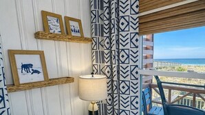 A room with a white desk, framed pictures, a lamp, patterned curtains, and a view of the beach.