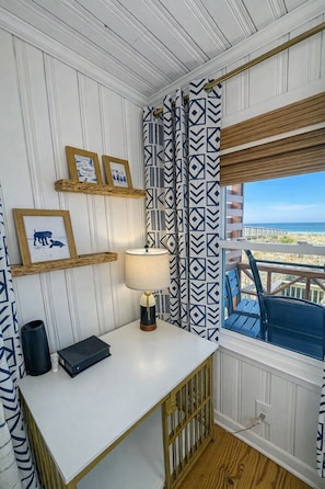 A room with a white desk, framed pictures, a lamp, patterned curtains, and a view of the beach.
