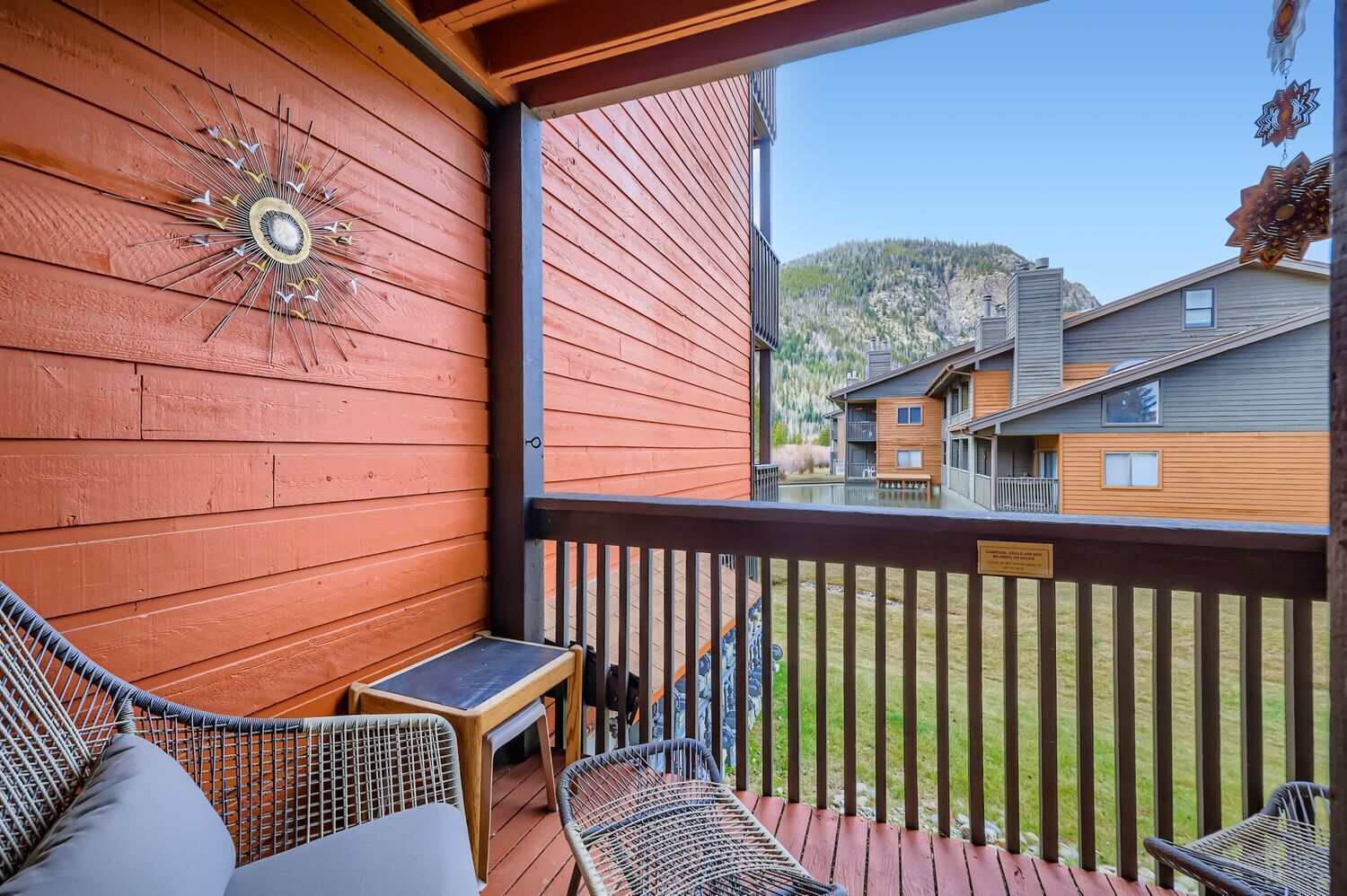 A wooden deck with wicker furniture, a decorative wall clock, and a view of other buildings and a mountain.