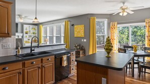 A modern kitchen with wooden cabinets, a black countertop, and a central island. There are two ceiling fans, a window with yellow curtains, and a dining area with a table and chairs. - The Yellow House - hot tub, pool table, fire pit, 6 bedrooms (Liberty)