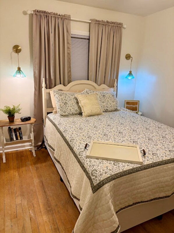 A bedroom with a bed, curtains, bedside lamps, a bedside table with a plant, and a framed picture.