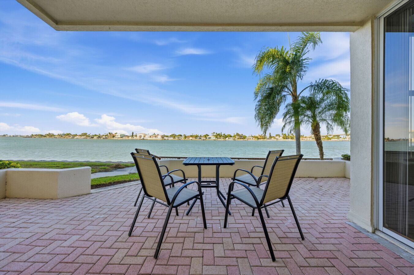 A patio with a table and chairs overlooking a body of water, palm trees, and a distant shoreline.