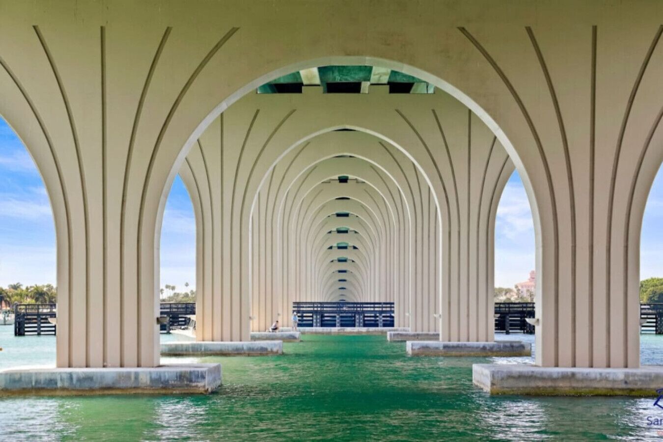 Un puente peatonal cubierto con una serie de arcos, un sendero verde y vistas al agua y edificios al fondo.