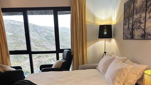 A hotel room with a large bed, a chair, a nightstand, and a view of a mountainous landscape through large windows. - Loft Eira Cálm&luxury Views Sierra Nevad Parking included (Sierra nevada)